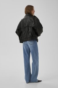 Vrouw met bruin haar, gekleed in een oversized zwarte leren jas, lichtblauwe wijduitlopende spijkerbroek en zwarte puntige schoenen, staand in zijwaartse houding.