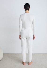 Vit långärmad topp med rund halsringning, tillsammans med strukturerade vita randiga byxor, barfota. Enkel, minimalistisk design och neutrala färger.
