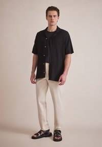Sort ribbet kortærmet skjorte, sort t-shirt underneath, lys beige bukser og sorte sandaler med remme. Enkel baggrund, neutrale nuancer.