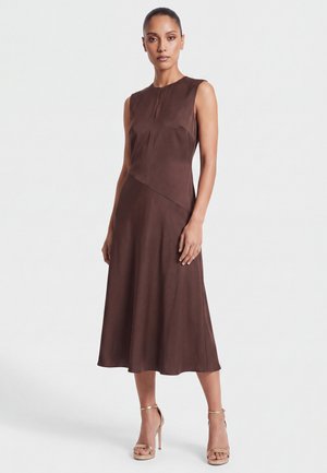 Woman standing wearing a sleeveless brown midi dress and beige high-heeled sandals against a plain white background.