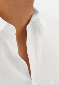 Camisa de lino blanca, con el cuello abierto, que presenta una sutil textura. Los botones son visibles, con un diseño limpio y minimalista, y una caída suave.