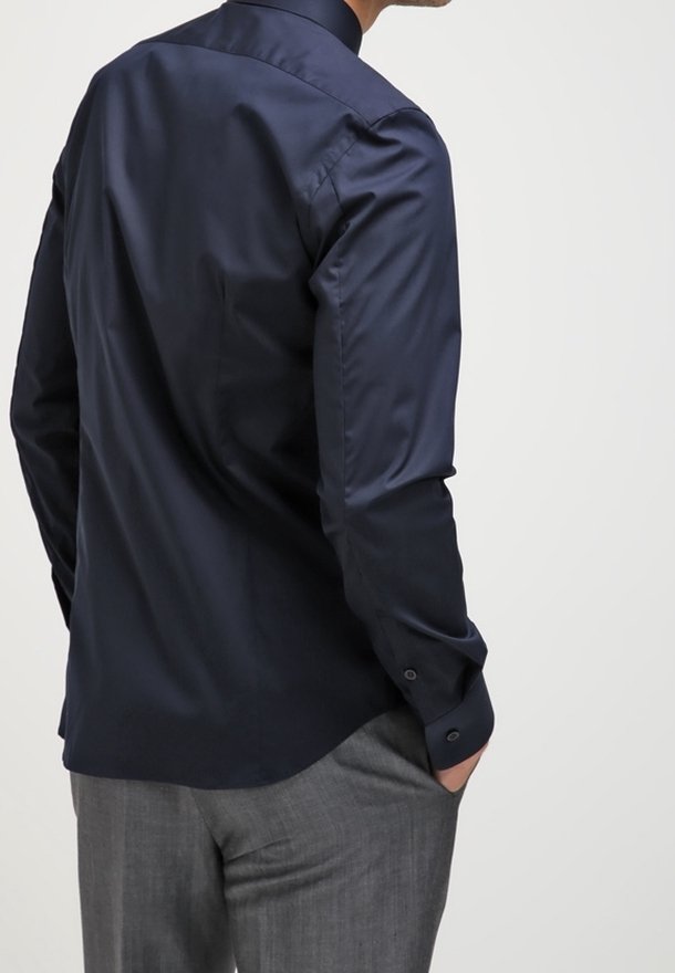 Homme portant une chemise manches longues bleu foncé et un pantalon gris, debout avec une main dans la poche, dos à la caméra sur un fond clair uni.