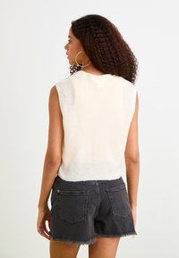 Maglione senza maniche color crema con scollo a coste, abbinato a pantaloni shorts in denim nero a vita alta con orlo sfrangiato e tasche posteriori.