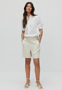 Witte button-up shirt met opgerolde mouwen, gecombineerd met beige shorts. De outfit heeft een schoon ontwerp en gladde texturen, met minimale details.