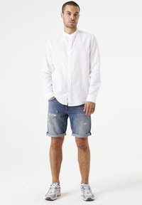 Camisa de lino blanca de mangas largas con cuello tipo bandana, combinada con shorts de denim desgastados. Los accesorios incluyen zapatillas deportivas grises.