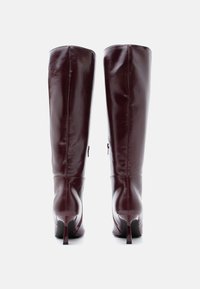 Botas de cuero burdeos hasta la rodilla con un acabado brillante, diseño elegante y tacones altos de aguja. Presentan un sutil detalle de cremallera en la parte trasera.