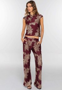 Mujer vistiendo un conjunto de top burdeos a juego y pantalones de pierna ancha con un patrón floral metálico, con las manos en los bolsillos, de pie frente a un fondo liso.