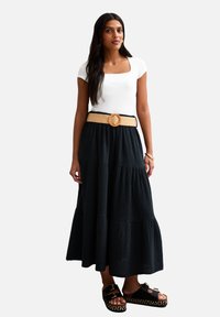 Gonna maxi nera a strati con tessuto testurizzato, abbinata a una maglietta bianca fitted e una cintura larga in paglia beige con fibbia rotonda. Sandali neri con plateau.