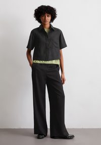 Geknipte zwarte blouse met een kraag en een borstzak, laagjes over een lichtgroene top. Losvallende zwarte broek en platte zwarte schoenen maken de look compleet.