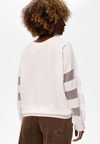 Crèmekleurige sweatshirt met doorschijnende gestreepte mouwen, ronde halslijn, ribbelboord onderaan en bij de mouwen. Gecombineerd met bruine flodderpantalon. Getextureerde stof.