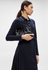 Tumši zila kleita ar garām piedurknēm, polo apkakli, baltu apdari un jostu viduklī. Melna, savākta rokassoma turēta vienā rokā.