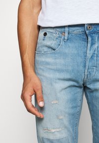 Světlé modré džíny z denimu s vybledlou texturou, s moderním poškozením v oblasti kolen a zapínáním na knoflík s rozepínáním na zip.