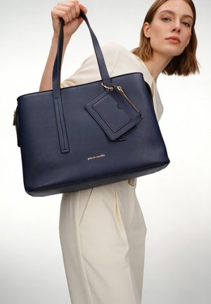 Vrouw in crèmekleurige outfit met grote marineblauwe leren tas met bijpassende kaarthouder en gouden ritssluitingdetails.