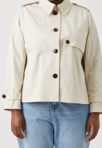 Veste cropped beige en tissu lisse, avec un col, des boutons foncés et des poches latérales. Jean en denim bleu clair visible.
