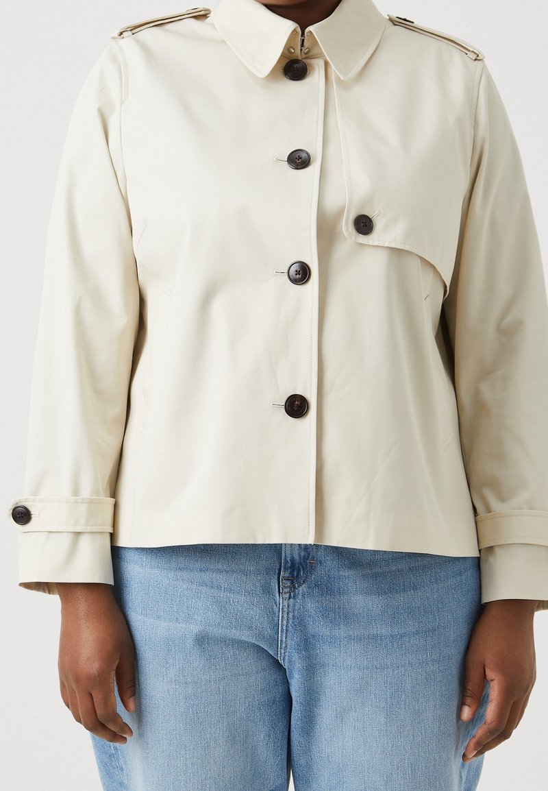 Veste cropped beige en tissu lisse, avec un col, des boutons foncés et des poches latérales. Jean en denim bleu clair visible.