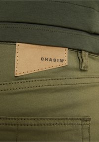 Olive green pants with a smooth texture, featuring a leather tag labeled "CHASIN'" attached to the waistband. Subtle stitching detail visible.