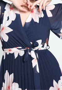 Femme portant une robe portefeuille bleu marine à fleurs avec une jupe plissée et une ceinture nouée à la taille, posant avec les mains près du visage.