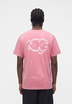 Mężczyzna z warkoczami cornrow nosi różową koszulkę Nike z dużym białym stylizowanym logo na plecach, w połączeniu z ciemnymi szortami.