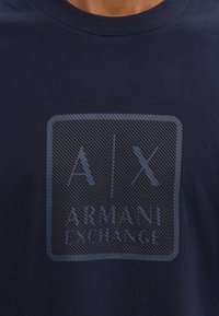 Camiseta de color azul marino hecha de algodón. Presenta un gráfico texturizado en azul oscuro con "A/X" y "ARMANI EXCHANGE" dentro de un contorno rectangular.