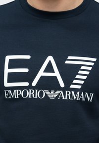 Mornarsko modra bombažna majica s kratkimi rokavi, na kateri je velik bel odtis logotipa: "EA7" in "EMPORIO ARMANI" z zgornjim črtastim poudarkom. Okrogel izrez ovratnika.