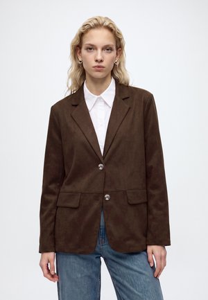 Femme aux cheveux blonds ondulés portant un blazer marron sur une chemise blanche et un jean bleu, debout sur un fond blanc uni.