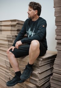 Schwarzes Langarmshirt mit Grafikdruck, schwarze Shorts und schwarze Wanderschuhe. Sitzt auf gestapelten Kartons.