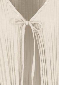 Light beige pleated textile featuring a tie closure at the neckline. The fabric appears soft with a subtle sheen and textured surface.