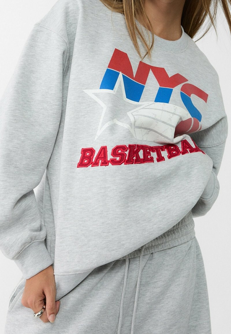 Harmaa collegepaita, joka on valmistettu pehmeästä materiaalista ja jossa on pyöreä kaula-aukko. Sisältää punaisia ja sinisiä grafiikoita, mukaan lukien "NYS" ja "BASKETBALL", sekä tähtimuotoilun.
