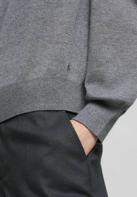 Maglione grigio lavorato a maglia con orlo e polsini a coste, caratterizzato da un piccolo dettaglio logo. Indossato con pantaloni scuri e texturizzati.