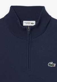 Pull en bleu marine zippé avec un col côtelé, présentant un petit logo crocodile vert de Lacoste sur la poitrine gauche. Tissu doux.