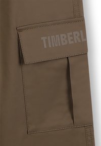Bruine stof met een gladde textuur, voorzien van een zijzak. Het woord "TIMBERL" is in lichtgrijs op de zakflap geperst.