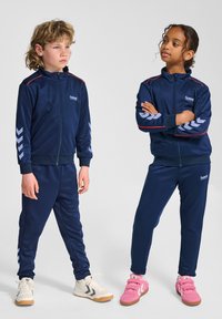 Navy Trackanzüge aus glattem Stoff mit roten Akzenten; einer mit weißen Chevron-Mustern an den Ärmeln. Junge in weißen Sneakers, Mädchen in pinken Schuhen.