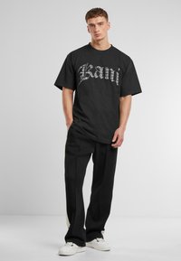 Black oversized T-shirt with bold silver graphic text, paired with black trousers and white sneakers, set against a light gray background.