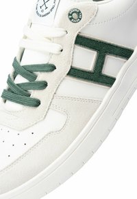 Zapatilla blanca con acentos verdes, materiales de ante y cuero, punta redonda, detalles perforados y cordones planos. Logotipo circular en la lengüeta.