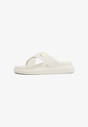 Sandales blanches à enfiler avec des lanières croisées, fabriquées en matériau lisse. Elles présentent une semelle légèrement texturée et un petit emblème doré sur la lanière.