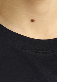 Camiseta de algodón negra con cuello redondo y tejido texturizado, con un lunar visible sobre un fondo de piel clara.