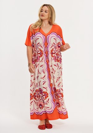 Frau in einem langen orange-cremefarbenen Blumenkleid mit lila Akzenten, die seitlich schaut, rote Ballerinas und goldene Armbänder trägt.