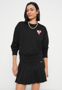 Μαύρο φούτερ με ροζ καρδιά, συνδυασμένο με μαύρη πλισέ φούστα. Υφασμάτινη υφή και ριμπ μανσέτες που αναδεικνύουν το σχέδιο.