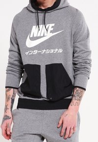 Šedá mikina s velkým bílým logem Nike a japonským textem. Má černé přední kapsy a lem s elastickou úpravou. Dlouhé rukávy jsou s manžetami.