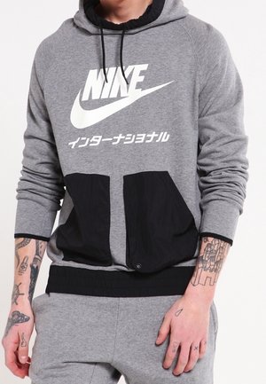Szara bluza z dużym białym logo Nike i japońskim tekstem. Posiada czarne kieszenie z przodu i elastyczny dół. Długie rękawy są wykończone mankietami.