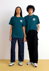 To personer har matchende kortermede t-skjorter i teal med et logo, kombinert med mørke jeans og sneakers. Enkelt design, avslappet passform.