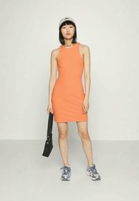 Vestido laranja justo, sem mangas, com um decote alto. Modelo usa ténis cinzas e segura uma mala preta, com um boné e um colar como acessórios.