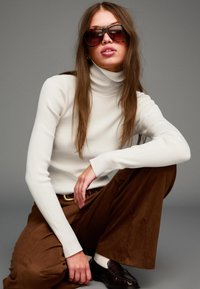 Weißer gerippter Rollkragenspullover, braune weiter geschnittene Hose, dunkle Sonnenbrille, braune Lederschuhe, glatte Texturen, minimales Design, neutrale Farben.