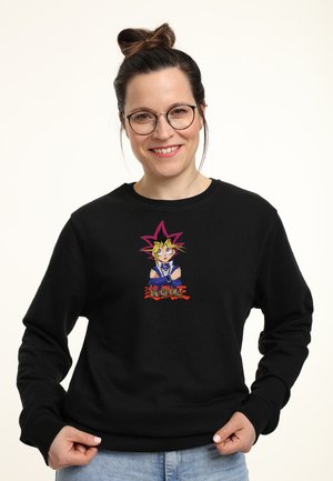 Kvinde med briller og håret samlet i en knold bærer en sort sweatshirt med farverig Yu-Gi-Oh!-figur og logo, smilende til kameraet.