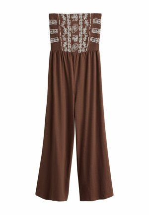 Vrouw in een strapless bruine jumpsuit met witte borduursels op het bovenstuk, zonnebril dragend, staand op een zanderig strand bij het water.