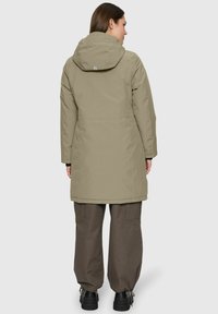Vrouw in een olijfgroene, knielange winterjas met capuchon, bruine losse broek en zwarte laarzen, van achteren gekeerd tegen een effen licht achtergrond.