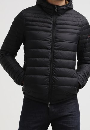 Mężczyzna w czarnej pikowanej kurtce puchowej z kapturem, zamkiem z przodu i rękami w bocznych kieszeniach, ubrany w ciemne dżinsy.