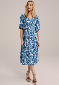 Blomstrede midi-kjole i blå nuancer med korte puffærmer, V-hals og bånd til at binde om taljen. Har et mønstret stof i nuancer af blå og hvid.