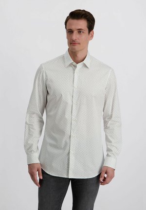 Mann, der nach vorne schaut, trägt ein weißes langärmliges Hemd mit kleinen schwarzen Polka-Dots und dunkelgraue Hosen vor einem einfarbigen Hintergrund.