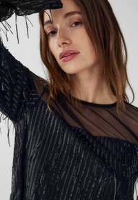 Czarny top z cekinami z przezroczystymi siateczkowymi rękawami, z pionowymi paskami i frędzlami, które tworzą teksturę i ruch.